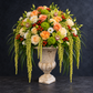 A large floral arrangement in a distressed stone urn vase, featuring peach and cream roses, white orchids, green mums, and cascading green trailing foliage, set against a dark textured background.