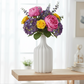A white ribbed ceramic vase holding a colorful flower bouquet with pink roses, yellow blooms, and purple hydrangeas, displayed on a light wood shelf in a bright, softly blurred room.