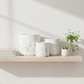 Three white ceramic planters with a gray marble pattern are arranged on a light wood floating shelf, alongside a small potted succulent in a glass vase and stacked books, against a warm off-white wall with soft diagonal shadows.