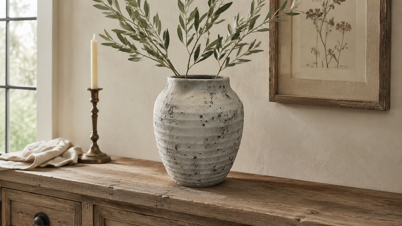 A textured, speckled gray ceramic vase filled with olive branches, displayed on a rustic wooden console table in a warm, neutral-toned room next to a candlestick and framed botanical art.