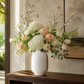A large floral arrangement in a textured white ceramic vase, featuring white hydrangeas, blush pink roses, and green foliage, displayed on a dark wood console table against a beige wall.