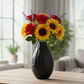 A black ribbed ceramic vase filled with a colorful floral arrangement of red roses, yellow sunflowers, and lavender sprigs, placed on a light wood table in a softly blurred living room setting.