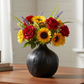 A black textured ceramic vase filled with a colorful flower arrangement featuring red roses, yellow sunflowers, and purple lavender sprigs, displayed on a wooden table in a bright indoor setting.