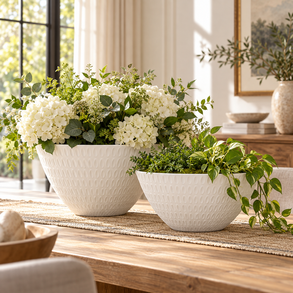 Two white textured ceramic planters filled with white flowers and green foliage, styled on a wooden table in a bright, modern living room.