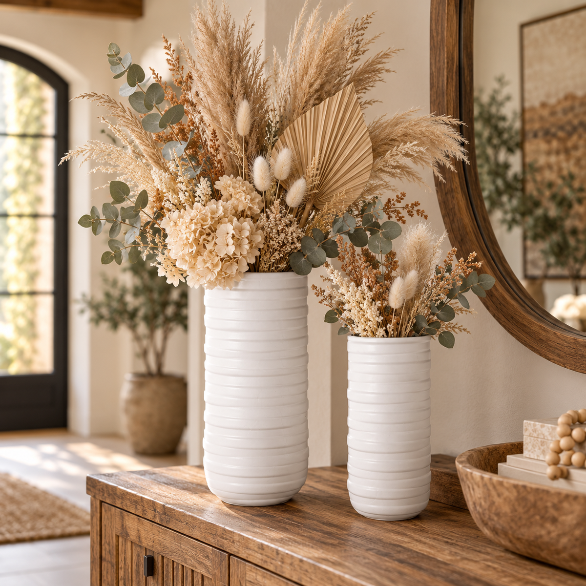 Two white ribbed ceramic vases filled with dried pampas grass, eucalyptus, and cream flowers, displayed on a wooden console table in a warm, styled interior.