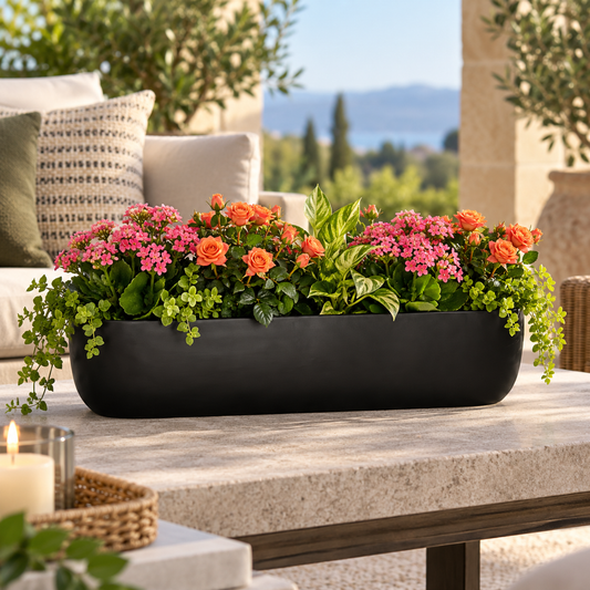 A long rectangular black planter filled with pink flowers, orange roses, and green foliage, placed on a stone patio table with an outdoor garden background.
