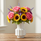 A vibrant floral arrangement featuring pink roses, yellow sunflowers, and purple lavender sprigs in a white ribbed ceramic vase, set on a rustic wooden table with a softly blurred window background.