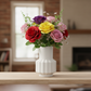 A colorful artificial rose bouquet in a white ribbed ceramic vase, displayed on a light wood table in a softly blurred living room setting.