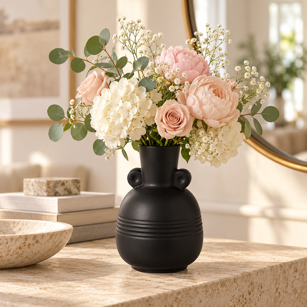 A matte black ceramic vase with a rounded body and two small side knobs, filled with a floral arrangement of blush pink and white flowers with green eucalyptus leaves, set on a stone countertop in a bright, modern interior.