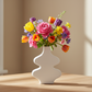 A white ceramic vase with a sculptural, wavy tiered design holding a vibrant bouquet of multicolored flowers, including pink roses, yellow tulips, and purple blossoms, set on a light wood table against a warm beige wall.