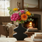 A black sculptural ceramic vase filled with a vibrant floral arrangement of pink, orange, and yellow flowers, set on a wooden table in a cozy living room with a blurred fireplace in the background.
