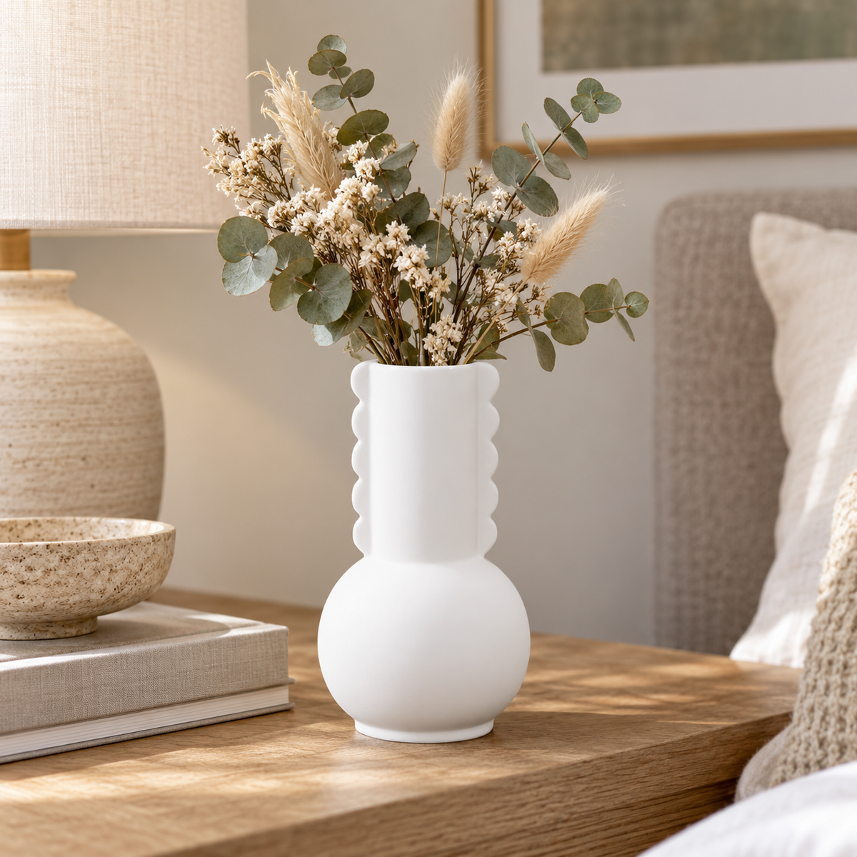 A white ceramic vase with a rounded body and scalloped neck detail, filled with dried grasses and eucalyptus stems, styled on a wooden bedside table next to a lamp and books.