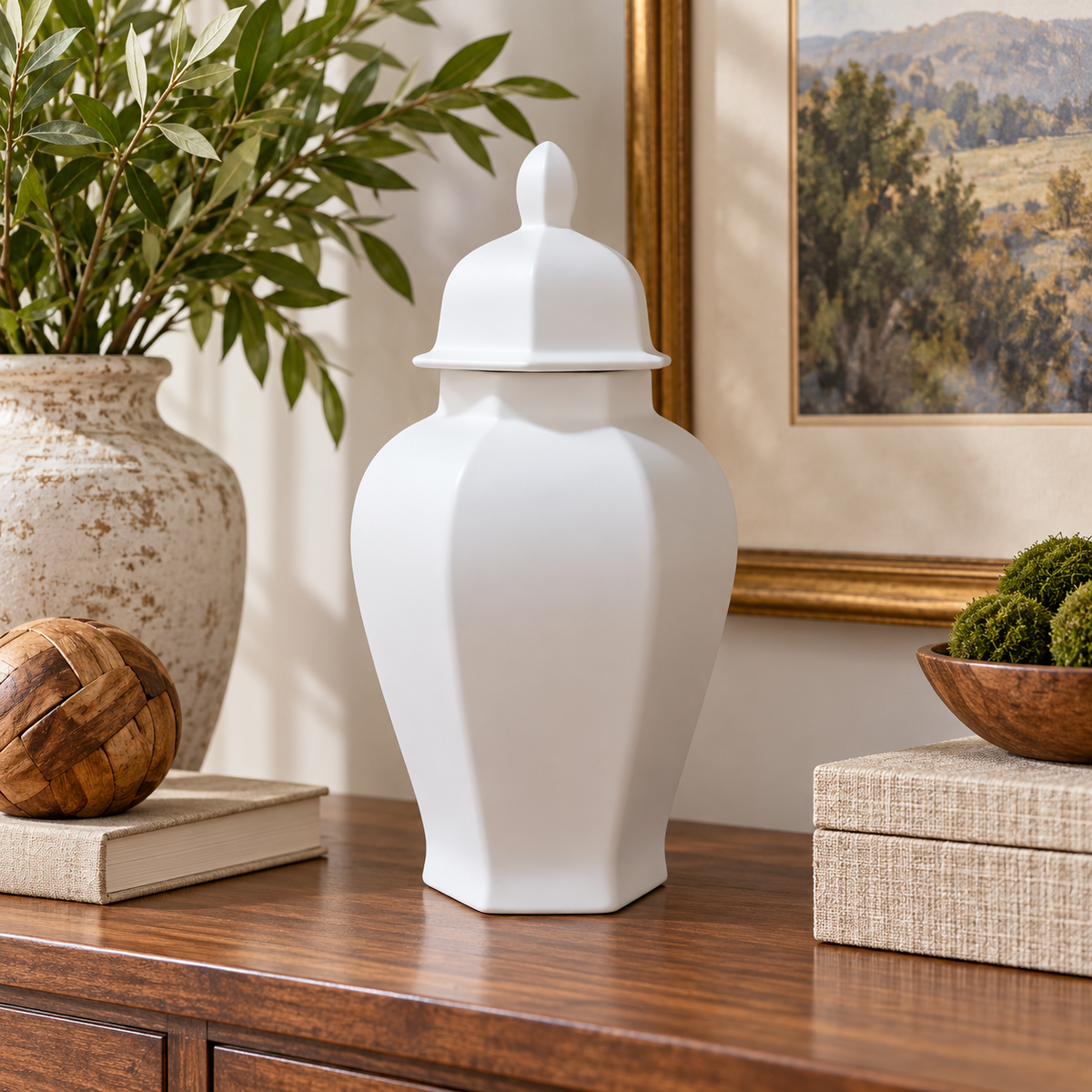 A white ceramic lidded storage jar with a flared base, displayed on a wooden console table in a styled home interior next to a potted plant and framed artwork.