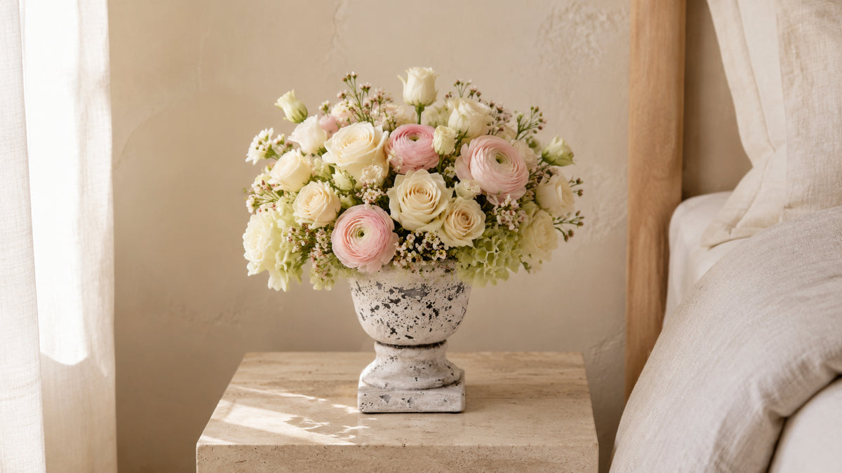 A floral arrangement of pale pink and cream flowers in a distressed white ceramic urn, displayed on a light wood bedside table next to a bed.