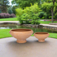 Two terracotta round flower pots placed on a surface with a garden and water body in the background.