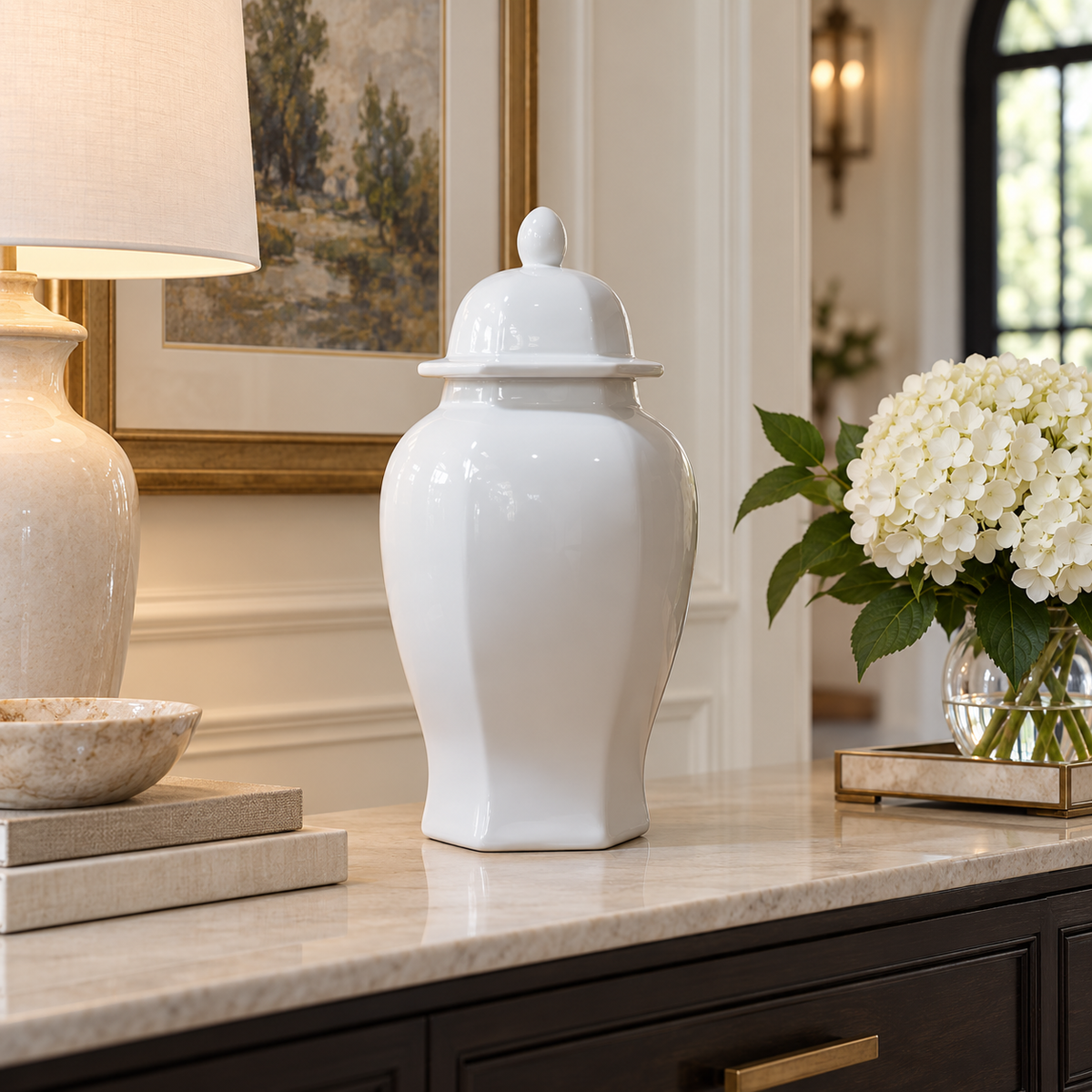 A glossy white ceramic ginger jar with a matching lid sits on a marble-topped console table in a beautifully decorated room. A lamp is on the left, and a vase of white hydrangeas is on the right.