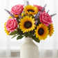 A vibrant artificial flower bouquet featuring pink roses, yellow sunflowers, and purple lavender sprigs arranged in a textured cream-colored ceramic vase, set on a light wooden surface against a soft, bright window background.