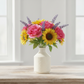 A vibrant artificial flower bouquet featuring pink roses, yellow sunflowers, and purple lavender sprigs, arranged in a textured cream-colored ceramic vase on a light wooden table.