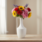 A white ceramic vase with a handle, filled with a colorful artificial flower bouquet featuring red roses, pink peonies, yellow sunflowers, and purple lavender, set on a light wooden table against a soft beige background.