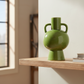 A glossy olive green ceramic vase with a bulbous body and two side handles, displayed on a light wood floating shelf in a modern interior setting.