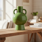 A glossy green ceramic vase with two side handles and a pedestal base, displayed on a wooden table in a softly lit, warm-toned interior setting.
