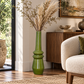 A tall, glossy green ceramic vase filled with dried grass stems, styled in a bright living room next to a wooden sideboard and a cream boucle armchair.