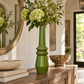 A tall glossy green ceramic vase filled with green hydrangeas and foliage, displayed on a wooden console table in a bright, modern interior setting.