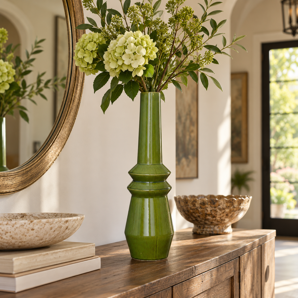 A tall glossy green ceramic vase filled with green hydrangeas and foliage, displayed on a wooden console table in a bright, modern interior setting.