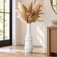 A tall white ceramic vase with a flared neck, filled with dried pampas grass and brown botanical stems, placed on a wood floor in a bright, modern room.