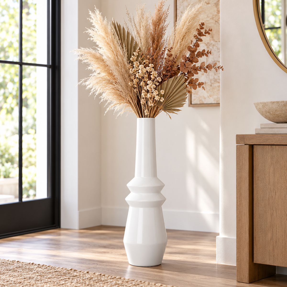 A tall white ceramic vase with a flared neck, filled with dried pampas grass and brown botanical stems, placed on a wood floor in a bright, modern room.
