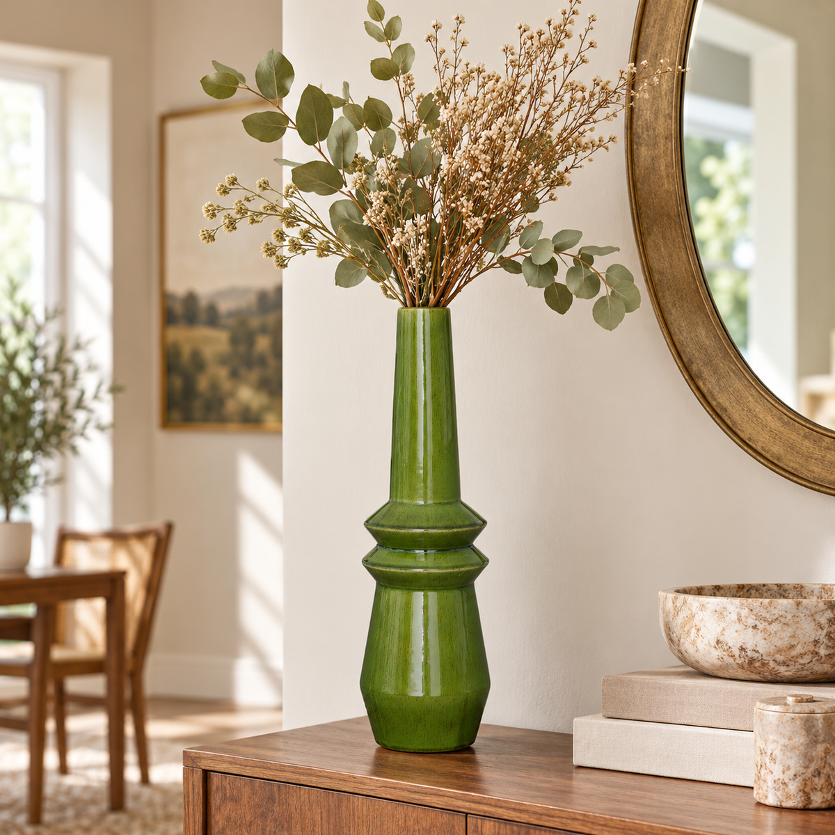 A tall glossy green ceramic vase filled with dried flowers and eucalyptus stems, styled on a wooden console table in a bright, modern living room with a round mirror and decorative bowls.