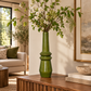 A tall glossy green ceramic vase filled with green leaves and small white flowers, displayed on a wooden console table in a styled living room interior.