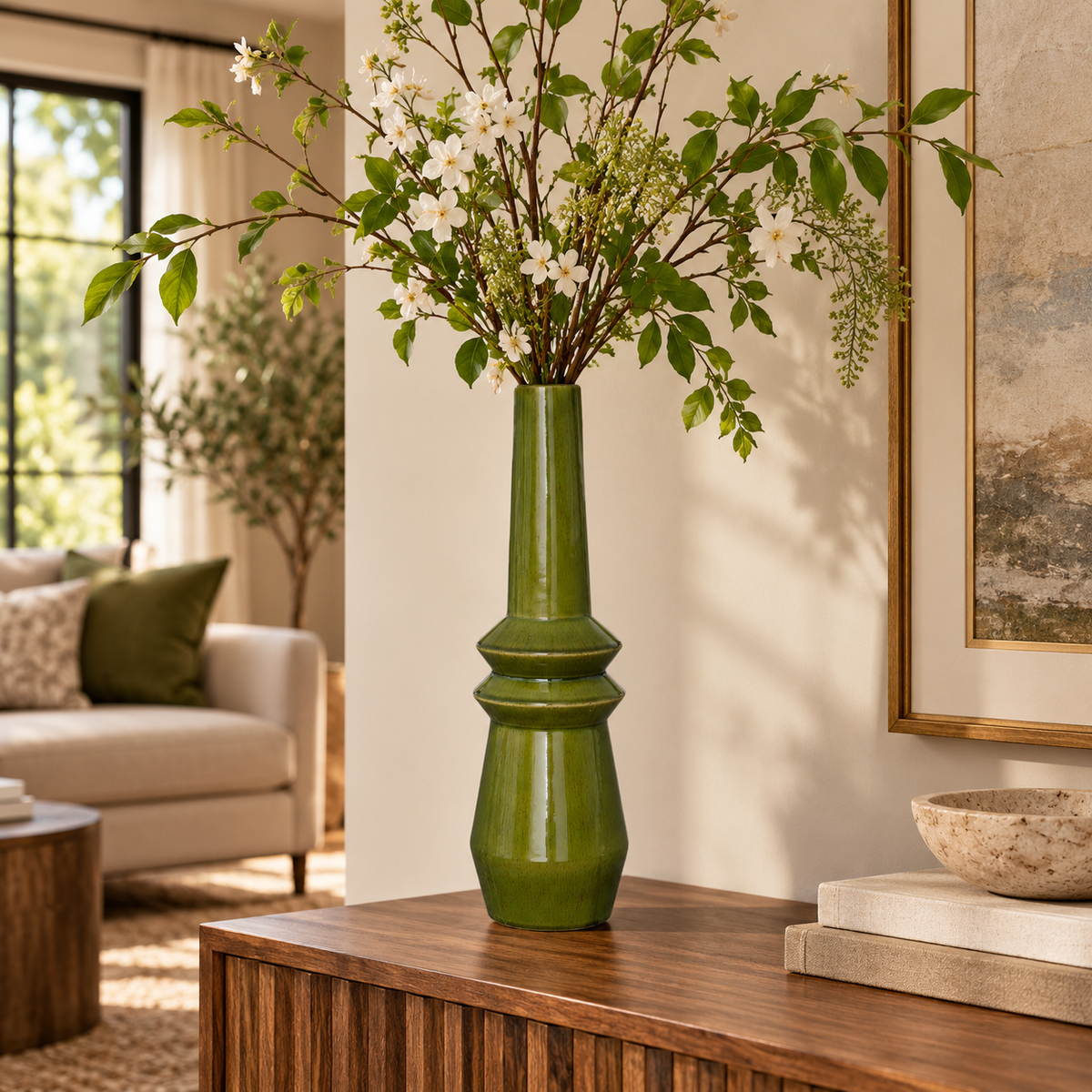 A tall glossy green ceramic vase filled with green leaves and small white flowers, displayed on a wooden console table in a styled living room interior.