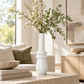 A tall, modern white ceramic vase with a flared base, filled with white blossoms and green eucalyptus stems, styled on a stone coffee table in a bright, contemporary living room.
