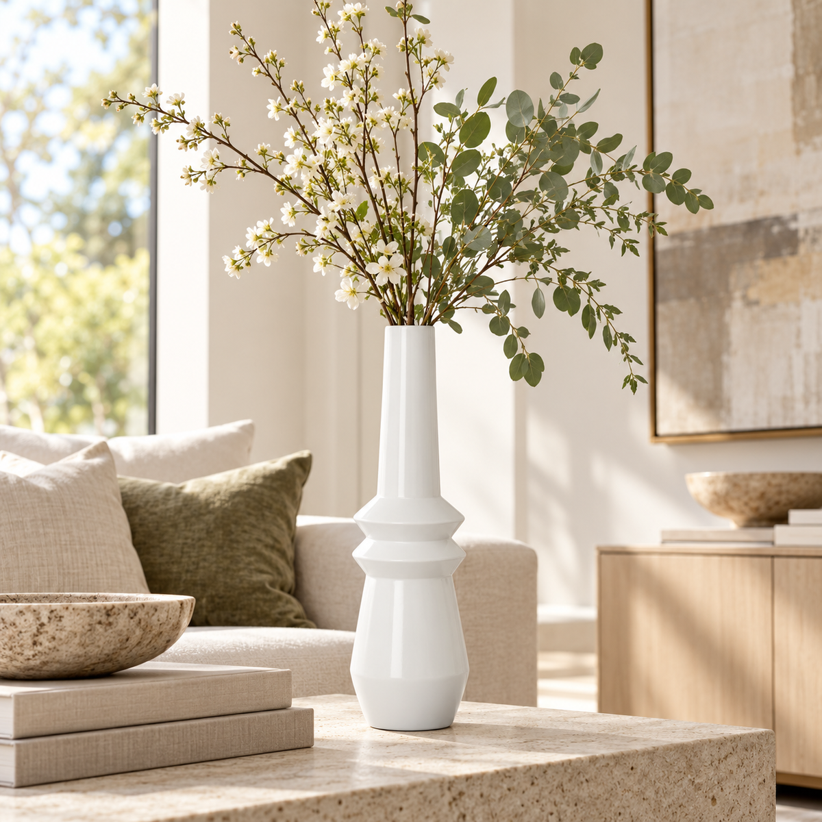 A tall, modern white ceramic vase with a flared base, filled with white blossoms and green eucalyptus stems, styled on a stone coffee table in a bright, contemporary living room.