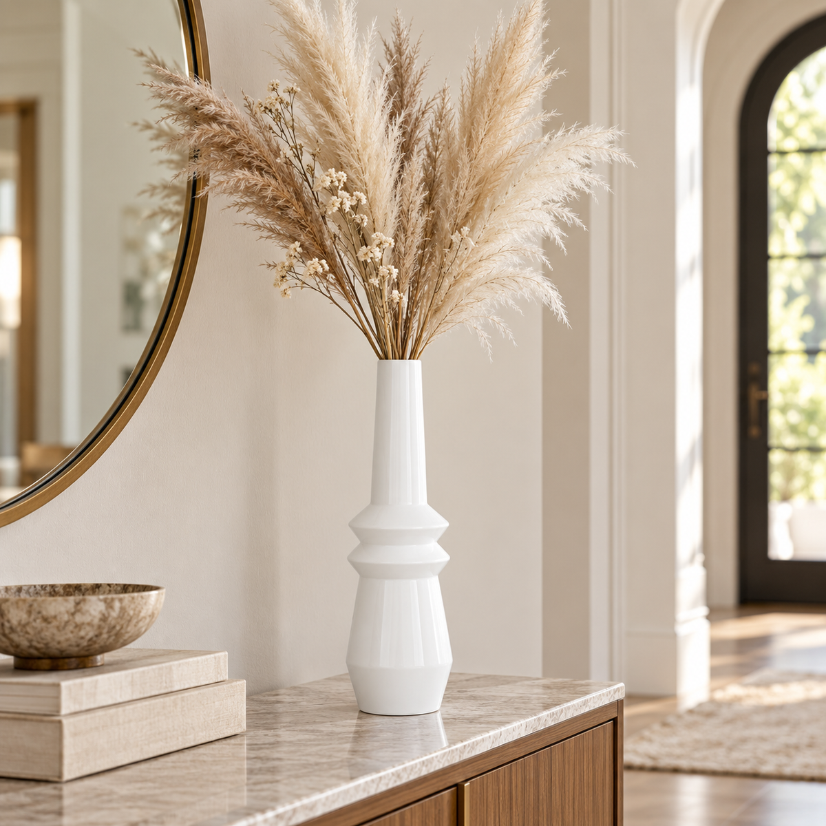 A tall white ceramic vase filled with beige pampas grass and small dried flowers, displayed on a marble-topped console table in a bright interior room.