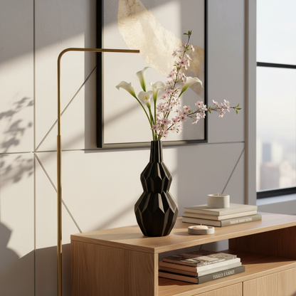 A black faceted vase holding white calla lilies and pink blossoms, styled on a light wood console table in a modern interior with a floor lamp, books, and framed wall art.