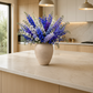 A large vase filled with a dense bouquet of blue, purple, and white flowers placed on a light marble kitchen island, with a modern kitchen in the background.