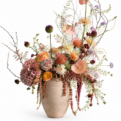 A rustic beige ceramic vase filled with a lush, multi-colored wildflower arrangement featuring orange ranunculus, pink anthuriums, purple globe flowers, and delicate trailing greenery, set against a solid white background.