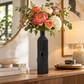 A black ribbed ceramic vase holding a colorful floral arrangement with pink, peach, white, and green flowers, displayed on a wooden console table in a warmly lit interior setting.
