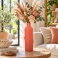 A coral pink ribbed ceramic vase filled with a dried floral arrangement, set on a wooden table in a bright, modern living room.