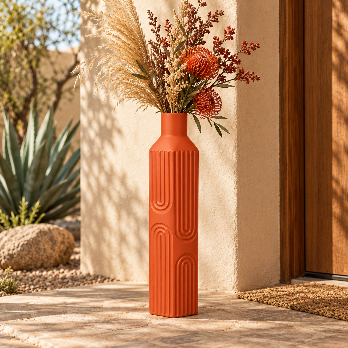 An orange terracotta ribbed ceramic vase filled with dried pampas grass and orange flowers, set on a stone patio in front of a beige stucco wall and wooden door.