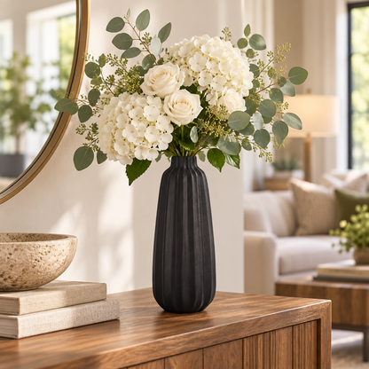 A black ribbed ceramic vase holding a white floral arrangement with hydrangeas, roses, and eucalyptus, displayed on a wooden console table in a bright living room.