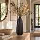 A ribbed matte black ceramic vase filled with dried ornamental grass and green leaves, displayed on a wooden console table in a warm, modern interior setting.