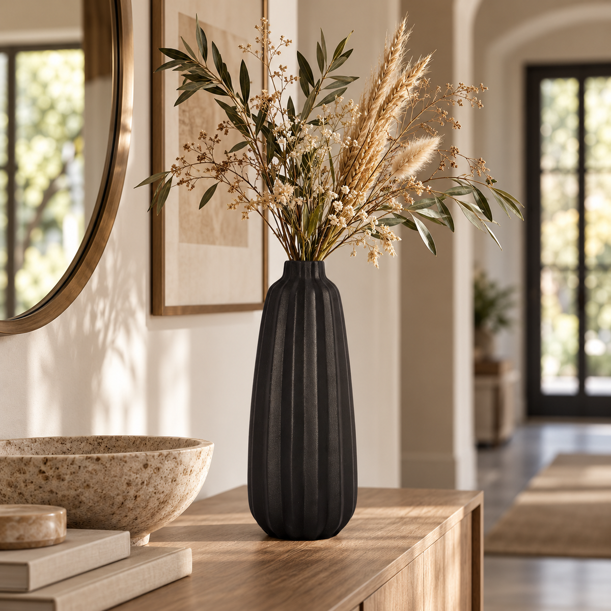 A ribbed matte black ceramic vase filled with dried ornamental grass and green leaves, displayed on a wooden console table in a warm, modern interior setting.