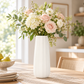 A tall white ribbed ceramic vase filled with a lush floral arrangement of blush pink roses, white hydrangeas, and green foliage, set on a wooden dining table in a bright, modern room.