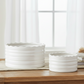 Two white ceramic planter pots with a scalloped rim and horizontal ribbed texture, displayed on a wooden table in a bright, softly lit room with sheer curtains, books, and a wooden bead garland.