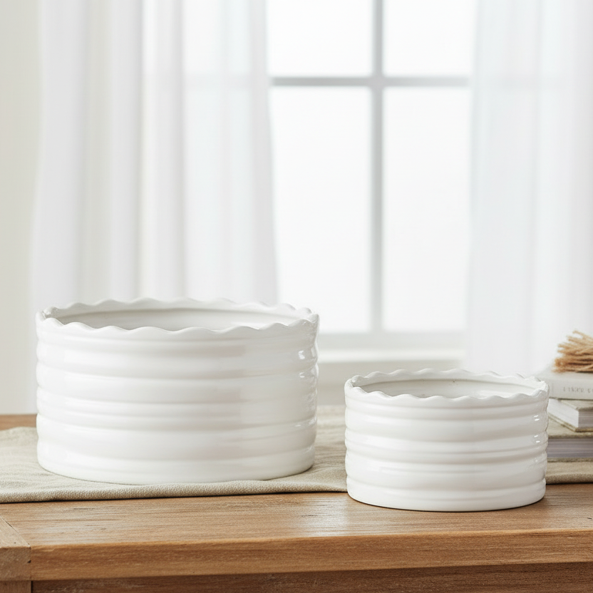 Two white ceramic planter pots with a scalloped rim and horizontal ribbed texture, displayed on a wooden table in a bright, softly lit room with sheer curtains, books, and a wooden bead garland.