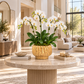 A potted white Phalaenopsis orchid arrangement in a textured gold ceramic pot, displayed on a round beige marble coffee table in a bright, modern living room.
