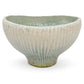 A handmade ceramic bowl with a ribbed, sunburst texture, featuring a glaze that transitions from a light cream-beige at the top to a textured light blue-green and earthy brown at the base, set against a plain white background.